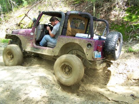1986 Ford Bronco - A purple Jeep Wrangler is driving through a muddy trail, with the driver visible behind the wheel. The vehicle is covered in mud, and water…