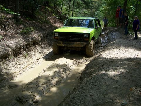 1986 Ford Bronco - A bright green 1986 Ford Bronco is navigating a deep mud rut on a wooded trail. Several people are standing on a higher bank watching the v…