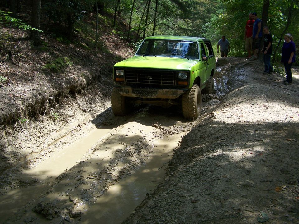 Green 1986 Ford Bronco driving through a muddy trail with people watching.
