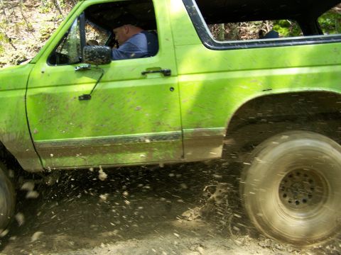 1986 Ford Bronco - A bright green Ford Bronco is driving through mud, with mud splashing up from the tires. The driver is visible behind the steering wheel.