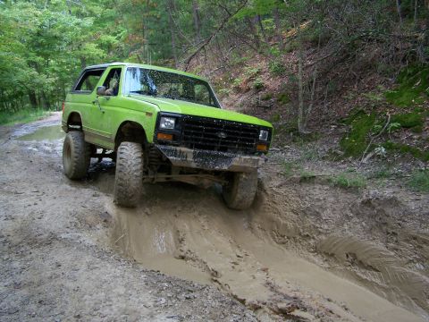1986 Ford Bronco - A bright green, lifted Ford Bronco is navigating a muddy, rutted trail in a wooded area. The vehicle is angled towards the viewer as it dri…