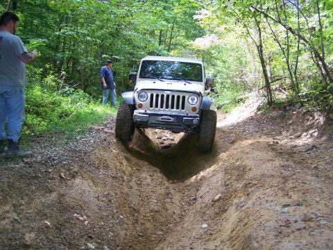 1986 Ford Bronco - A white Jeep Wrangler is navigating a deep rutted dirt trail in a wooded area. Two people are visible standing on the side of the trail.