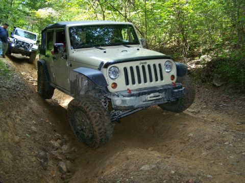 1986 Ford Bronco - A tan Jeep Wrangler is navigating a steep, muddy off-road trail. Another white Jeep is visible further up the trail.