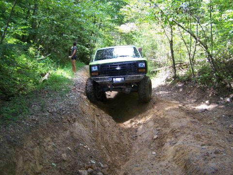 1986 Ford Bronco - A lifted, green Ford Bronco is driving down a muddy, rutted dirt trail in a wooded area. A woman stands on the side of the trail watching t…