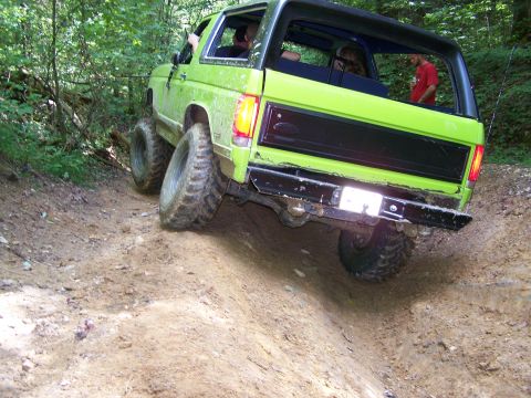1986 Ford Bronco - A bright green 1986 Ford Bronco is shown from the rear-side angle as it navigates a steep dirt trail in a wooded area. The vehicle has larg…