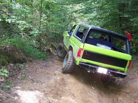 1986 Ford Bronco - A bright green Ford Bronco is driving off-road on a dirt path through a wooded area. The vehicle is angled up a hill with its rear facing t…
