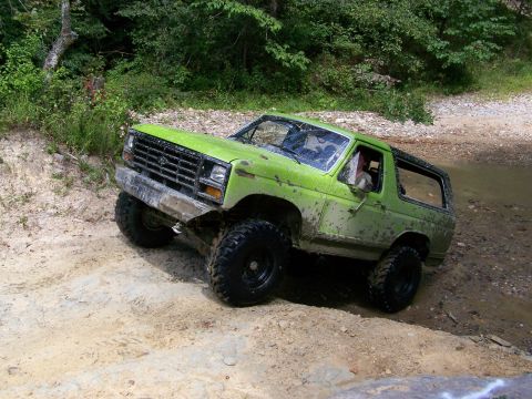 1986 Ford Bronco - A bright green, mud-covered 1986 Ford Bronco is navigating a muddy, off-road trail with steep inclines. The vehicle is tilted significantly…