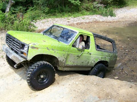 1986 Ford Bronco - A lime green Ford Bronco is stuck in a muddy ditch off-road. The vehicle is covered in mud, and the front tire is submerged in water.