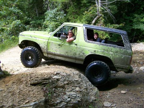 1986 Ford Bronco - A muddy, lime green 1986 Ford Bronco is navigating a rocky, off-road trail with two people inside. The vehicle is tilted to the side as it…