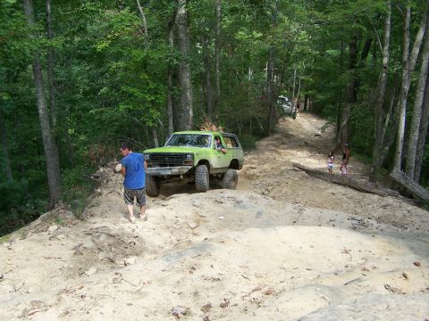 1986 Ford Bronco - A lime green 1986 Ford Bronco is navigating a steep, sandy off-road trail surrounded by dense forest. A man in a blue shirt stands on the s…