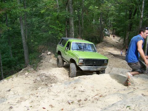 1986 Ford Bronco - A bright green Ford Bronco is navigating a steep, sandy off-road trail in a wooded area. A man in a blue shirt and plaid shorts is walking…