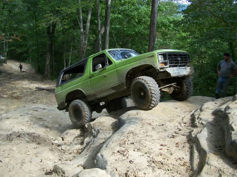 1986 Ford Bronco - A lifted green 1980s Ford Bronco is navigating a rocky, off-road trail in a wooded area. A man stands to the right of the vehicle, observin…