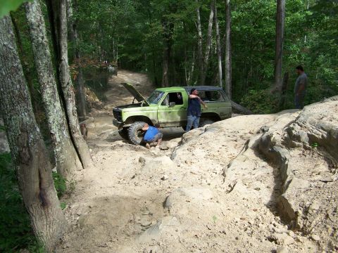 1986 Ford Bronco - A light green 1986 Ford Bronco is parked on a dirt trail in a wooded area with the hood open. Two men are standing near the vehicle, and an…