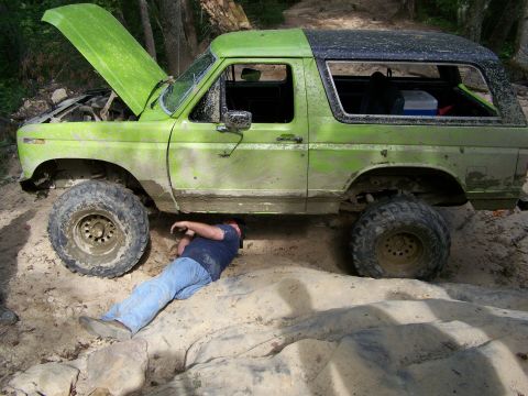 1986 Ford Bronco - A person lies underneath a muddy, lime green 1980s Ford Bronco with the hood up, appearing to work on the vehicle. The Bronco is on a dirt…