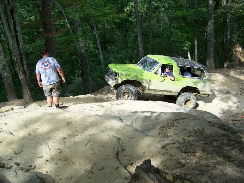 1986 Ford Bronco - A green 1986 Ford Bronco is shown off-roading on a dirt trail with a person standing nearby. The driver is visible inside the vehicle.