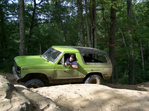 1986 Ford Bronco - A muddy, lime green 1980s Ford Bronco is stuck on a dirt trail in a wooded area. The driver is visible through the open driver's side windo…