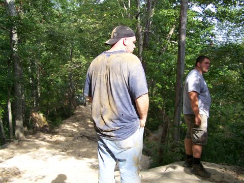 1986 Ford Bronco - Two men are standing on a dirt path in a wooded area. The man in the foreground is wearing a dirty t-shirt and jeans, and the man in the ba…