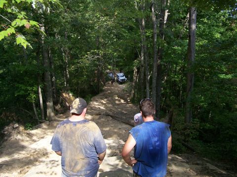 1986 Ford Bronco - Two men stand on a dirt path in a wooded area, looking down the path where several off-road vehicles are visible. The men are wearing dirty…