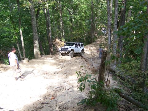 1986 Ford Bronco - A white Jeep Wrangler is parked on a dirt trail in a wooded area, with a man standing to its left and another man walking up the trail behi…