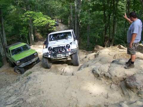 1986 Ford Bronco - A green 1986 Ford Bronco is on a steep, rocky trail, tilted to its left side. A white Jeep is behind it, and a man in a grey t-shirt and ca…