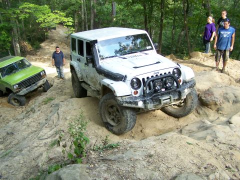 1986 Ford Bronco - A white Jeep Wrangler is navigating a steep, rocky, off-road trail. A green off-road vehicle is visible on the left side of the trail.