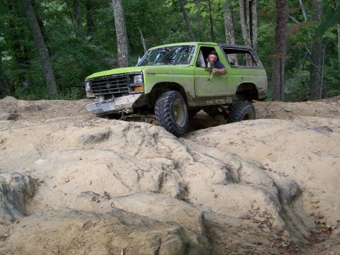 1986 Ford Bronco - A green 1980s Ford Bronco is navigating a rocky, off-road trail in a wooded area. The driver is visible behind the wheel, looking out the w…