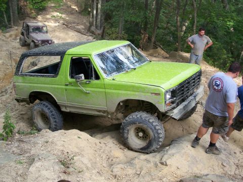 1986 Ford Bronco - A bright green Ford Bronco is navigating a steep, rocky trail with a purple Jeep visible further up the path. Two men are standing nearby,…