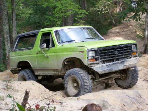 1986 Ford Bronco - A lime green, two-door Ford Bronco is navigating an off-road trail with large tires and a lifted suspension. The vehicle is covered in mud,…