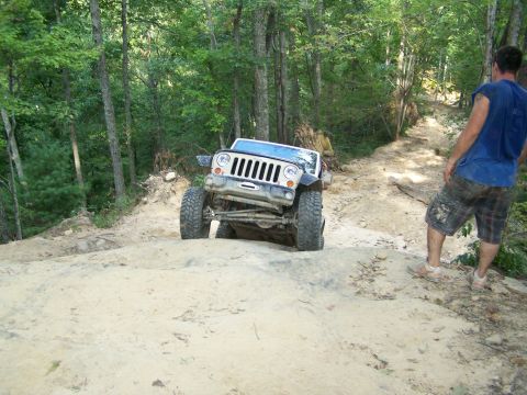 1986 Ford Bronco - A tan Jeep Wrangler is navigating a steep, sandy incline on a wooded trail. A man in a blue shirt and plaid shorts stands nearby, observing…