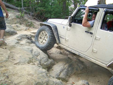 1986 Ford Bronco - A white Jeep Wrangler is navigating a rocky, off-road trail with a person walking alongside it. The Jeep's tires are large and aggressive,…