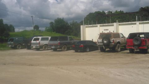 1996 Ford Bronco - Several Ford Broncos, SUVs and a lifted truck parked in a gravel lot beside a concrete wall under overcast skies.