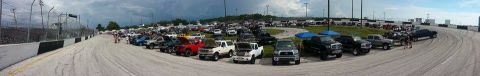 1995 Ford Bronco - A wide-angle view captures a gathering of trucks and SUVs at a race track event, under a cloudy sky.