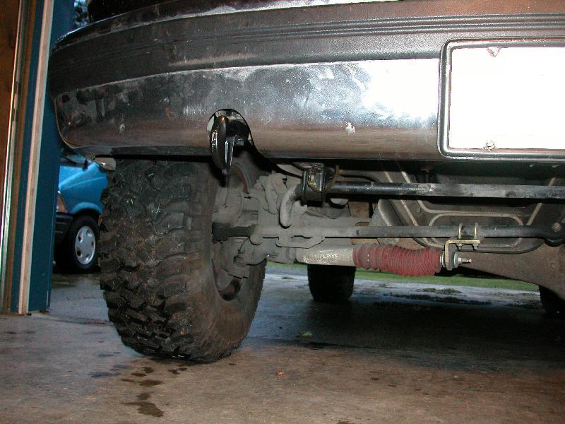 Underside front view of dark Bronco showing solid axle, mud tire and tow hook in a garage
