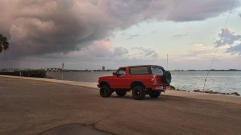 1995 Ford Bronco - A red 1995 Ford Bronco is parked near the water under a dramatic, cloudy sky.
