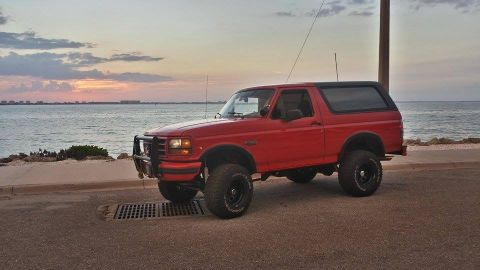 1995 Ford Bronco - A red Ford Bronco sits parked near the ocean as the sun sets in the background.