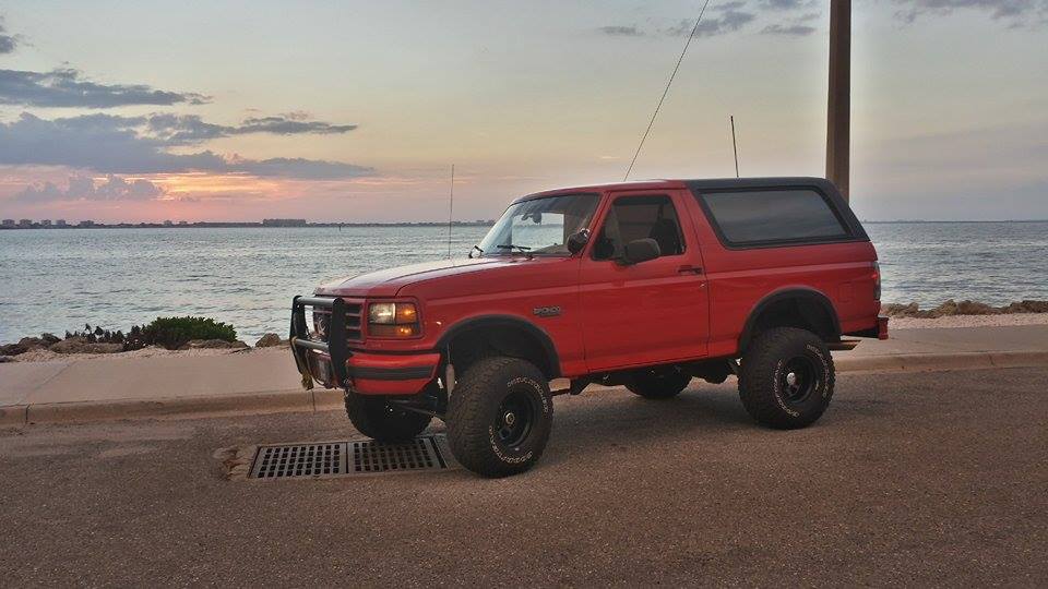 Red Ford Bronco parked by the water at sunset. It has a bull bar on the front