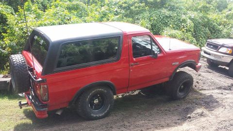 1995 Ford Bronco - A red Ford Bronco with a black top is parked next to another SUV on a dirt lot.