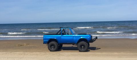 1974 Ford Bronco - A blue 1974 Ford Bronco is parked on a beach with the ocean waves in the background.