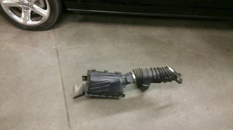 2004 Ford Crown Victoria - An air intake assembly is shown on a concrete floor, with the wheel and lower body panel of a black car visible in the background.