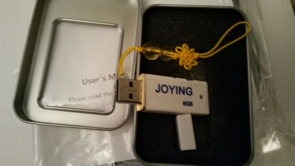 White USB flash drive labeled "JOYING 8GB" with a yellow cord and charm, inside a metal case with a user manual