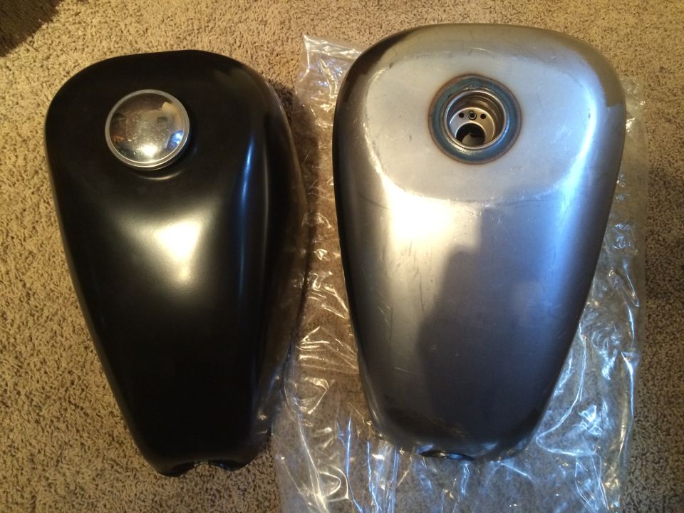 Two motorcycle fuel tanks, one black with a chrome cap, the other unpainted metal with a fuel cap opening