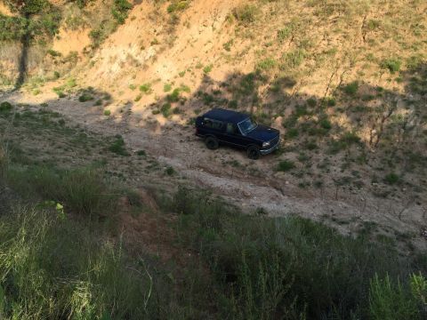 1993 Ford Bronco - A dark SUV drives on a dirt path through a dry, hilly landscape.