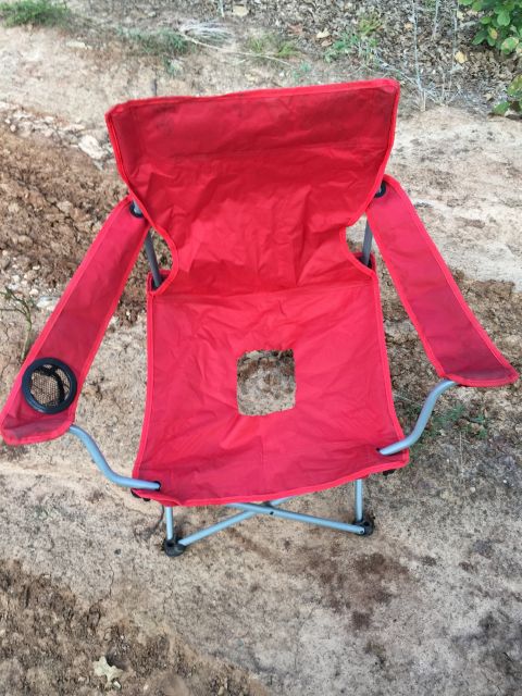 1993 Ford Bronco - A red folding camping chair with a square cutout in the seat sits on the ground. The chair has metal legs and armrests with built-in cup ho…