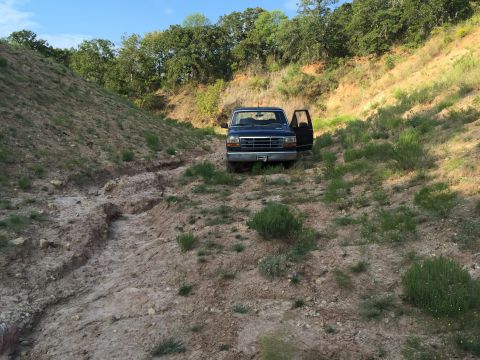 1993 Ford Bronco - A dark blue pickup truck is parked on a dirt path with its driver's door open, surrounded by dry vegetation and hills.