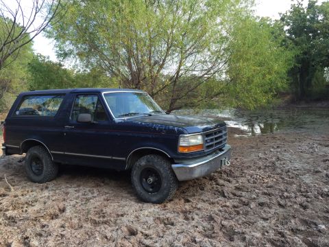 1993 Ford Bronco - A dark blue Ford Bronco is parked on a muddy bank next to a body of water with lily pads. The vehicle is angled towards the right, showing…
