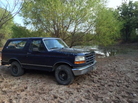 1993 Ford Bronco - A dark blue Ford Bronco is parked on a muddy bank next to a body of water with trees in the background. The SUV is angled towards the viewe…