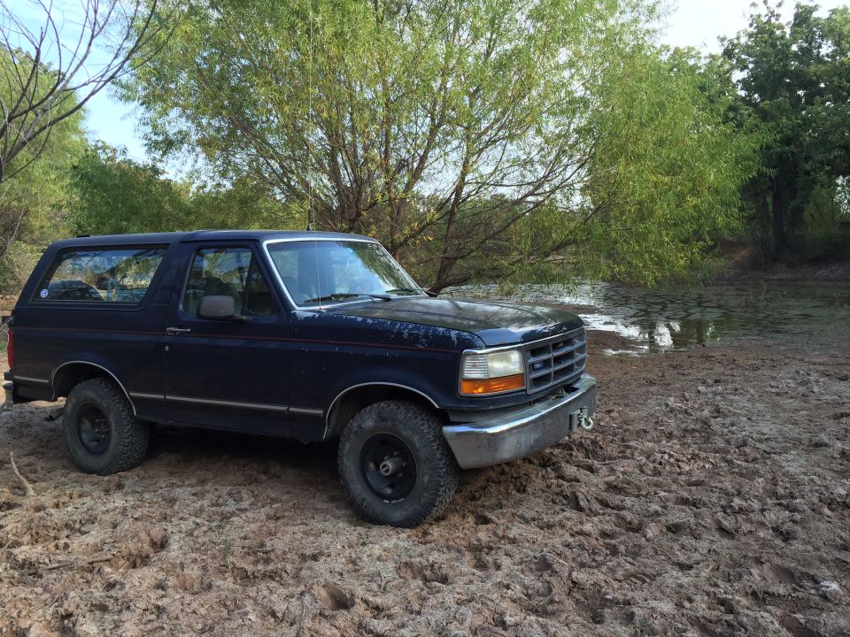 Dark blue 1993 Ford Bronco parked on muddy bank next to water.