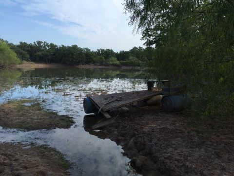 1993 Ford Bronco - A homemade raft made of barrels and wood sits on the muddy bank of a pond, partially submerged in the water. Trees and vegetation surround…