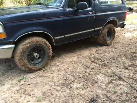 1993 Ford Bronco - A dark blue Ford Bronco is stuck in thick mud, with its front tire deeply embedded. The vehicle's body shows some wear and tear, and the ti…