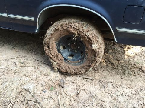 1993 Ford Bronco - A dark blue Ford Bronco's front wheel is deeply stuck in thick mud, with the tire completely caked and obscured by the wet earth. The vehic…